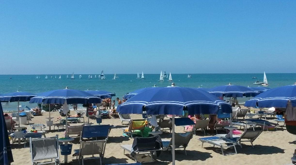 Stabilimento balneare con ombrelloni e lettini a disposizione dei clienti. Sul mare in lontananza ci sono numerose barche a vela.