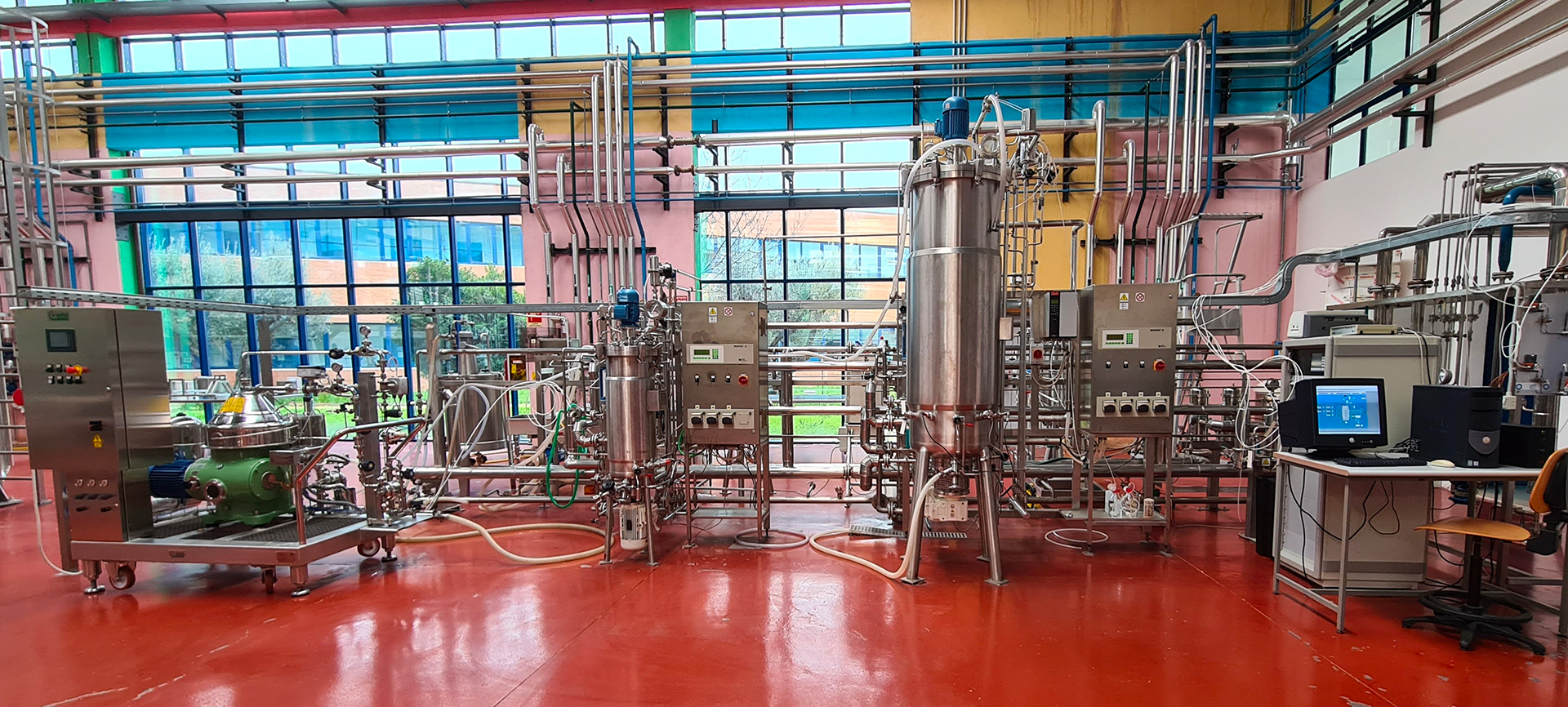 L’immagine mostra un impianto pilota per processi biotecnologici. Si vedono diverse apparecchiature in acciaio inox collegate da tubazioni e valvole, tra cui vessel da 50 e 500 litri e pannelli di controllo. A destra è presente una postazione con computer e monitor per il controllo del processo.